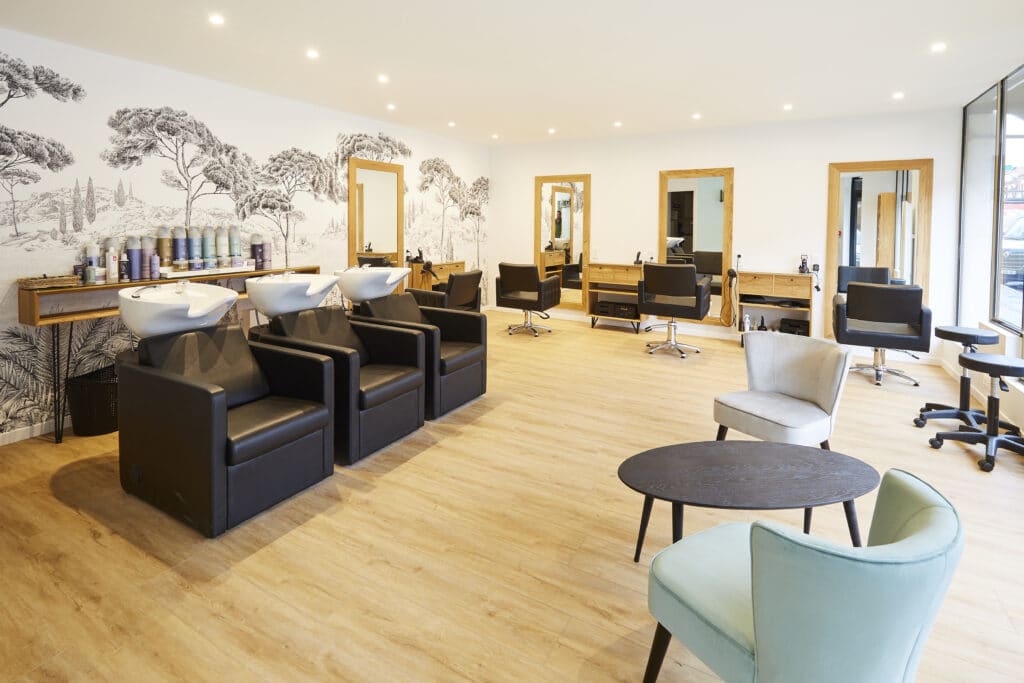 Salon de coiffure spacieux et moderne réalisé sur mesure par Kopocreation ébénisterie, avec fauteuils de lavage noirs devant mur décor paysage noir et blanc, postes de coiffure alignés à miroirs bois, sol en bois clair, éclairage LED, zone d'attente confortable avec fauteuils bleu clair et beige et table ronde noire