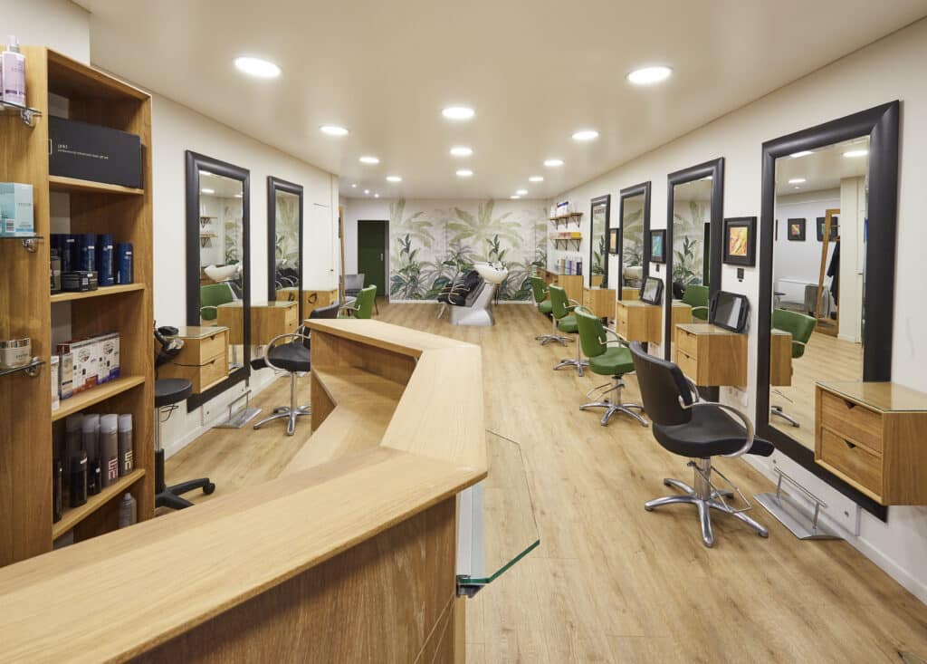 Salon de coiffure moderne aménagé sur mesure par Kopocreation ébénisterie, comptoir d'accueil en bois clair, postes de coiffure avec miroirs noirs, fauteuils verts, meubles en bois, sol bois clair, éclairage LED, zone de lavage au fond avec fauteuils et mur à motifs feuilles tropicales